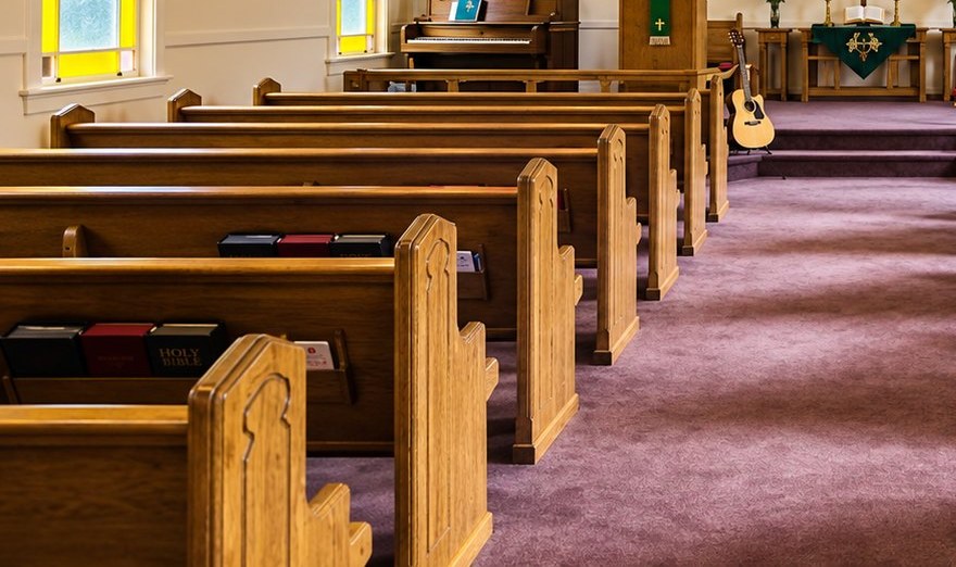 Oak pews at Blackburn Church, hymnals and Bibles in the racks, looking down the centre aisle over purple carpet.